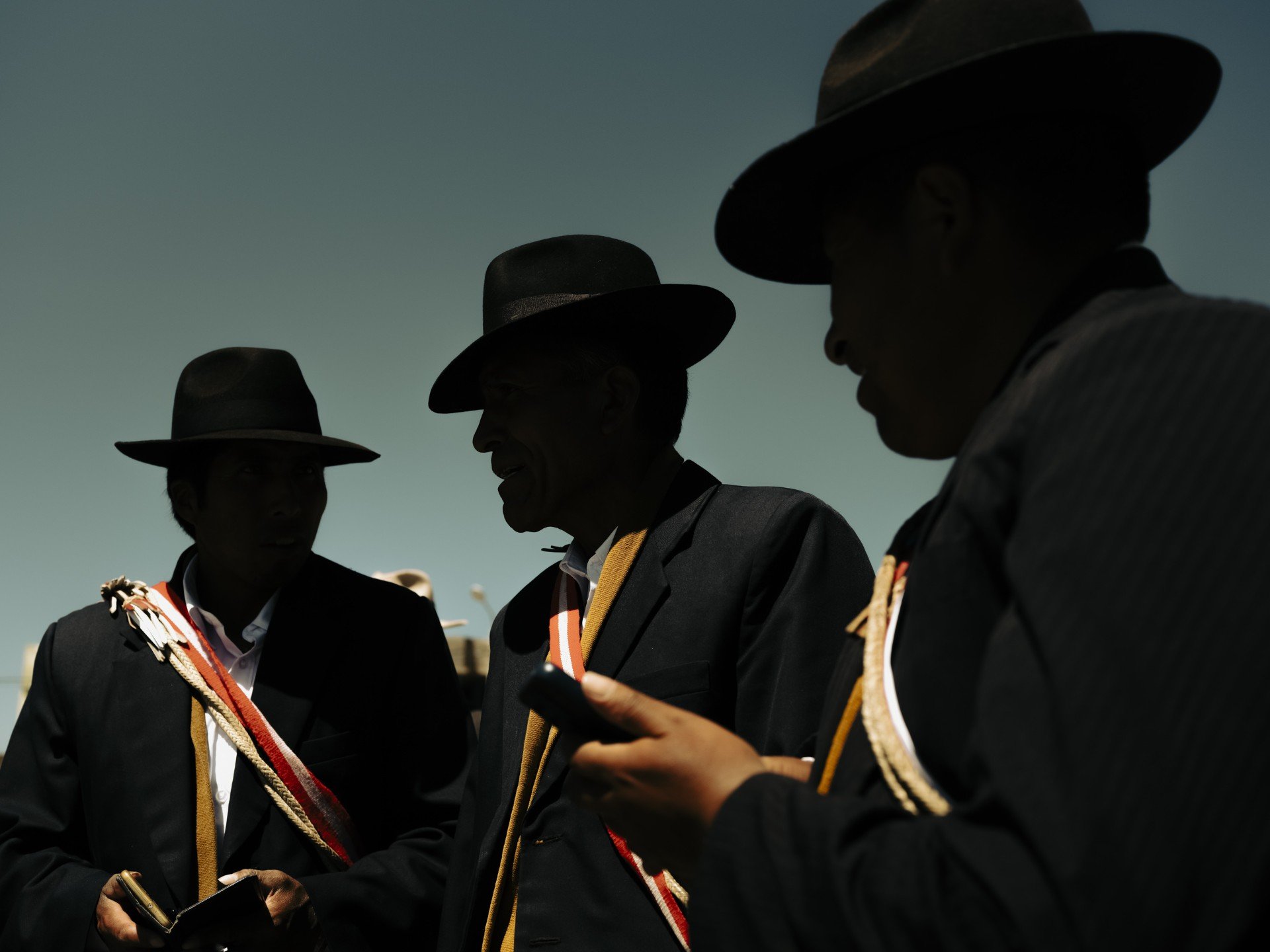 Siluetas oscuras de tres hombres (posiblemente autoridades o lancheros) con sombreros, conversando y mirando un teléfono contra un cielo dramático.